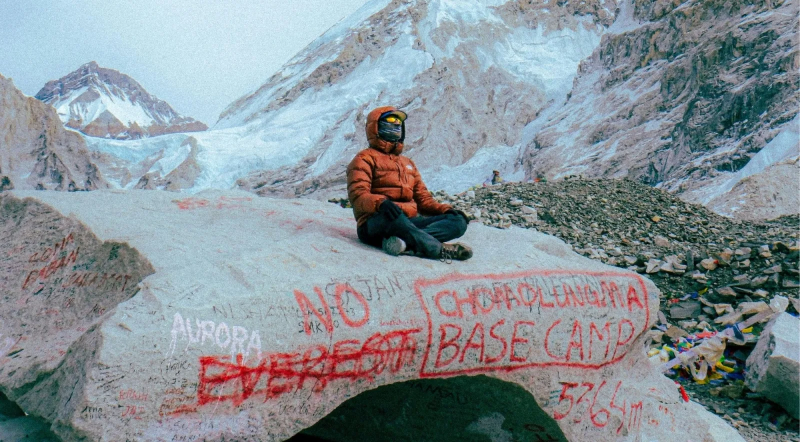 Trekker seated on the rock inscribed Everest Base Camp 5364m, prayer flags beside
