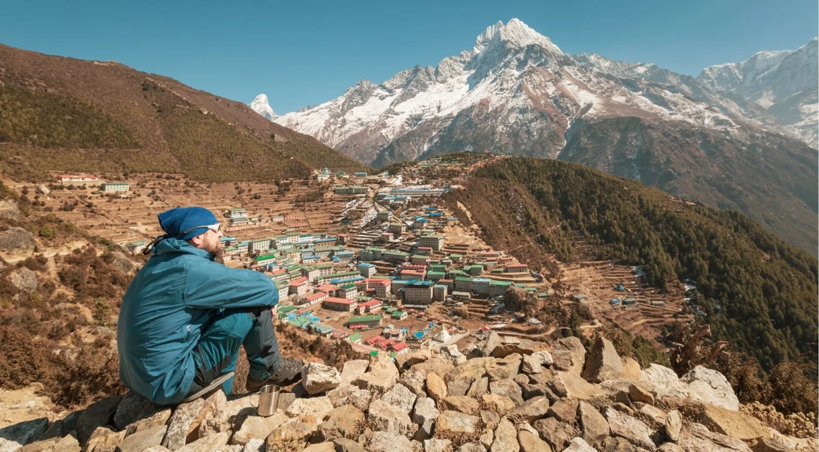 Trekker resting on a stone wall overlooking Namche Bazaar with snow peaks behind